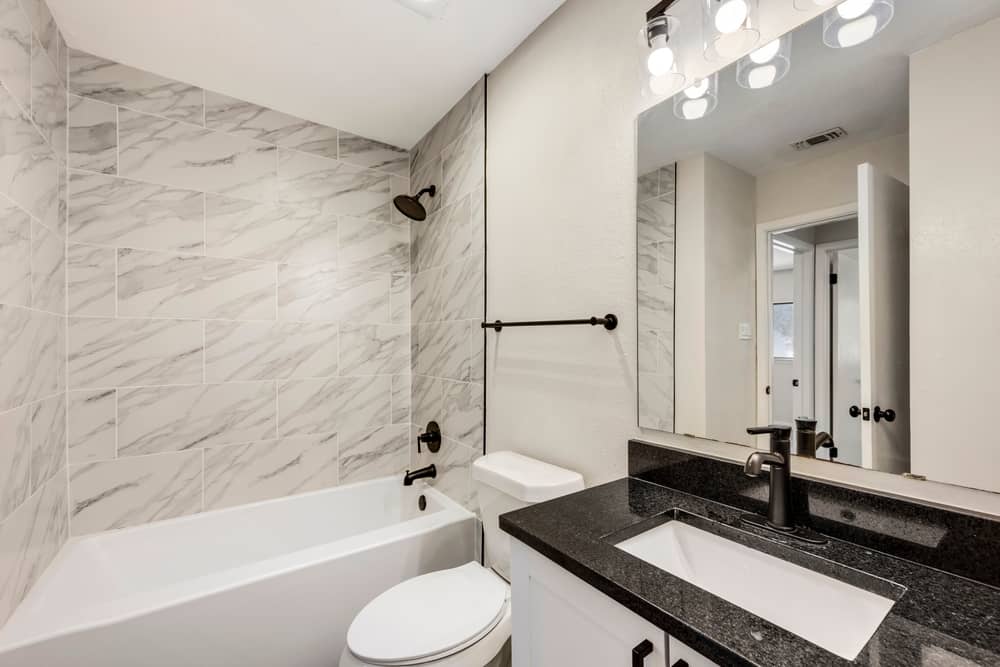 A modern bathroom with a white bathtub featuring marble tiles on the wall. The vanity has a black granite countertop with a white sink, black faucet, and a large mirror above, illuminated by several light bulbs. For expert Remodeling Services in Orlando Florida, this decor includes black fixtures and a towel bar.