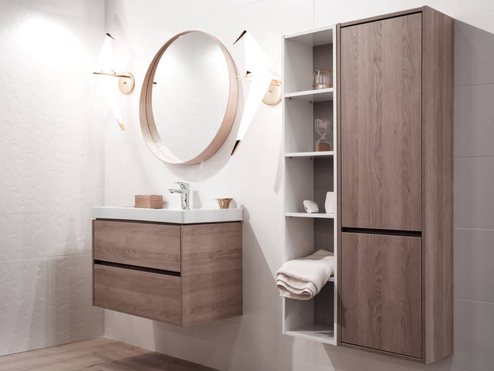 Modern bathroom with wood cabinets and a circular mirror. The vanity has two drawers, and the cabinet is open with towels and toiletries. The walls are white with subtle texture, and the lighting is soft and warm.