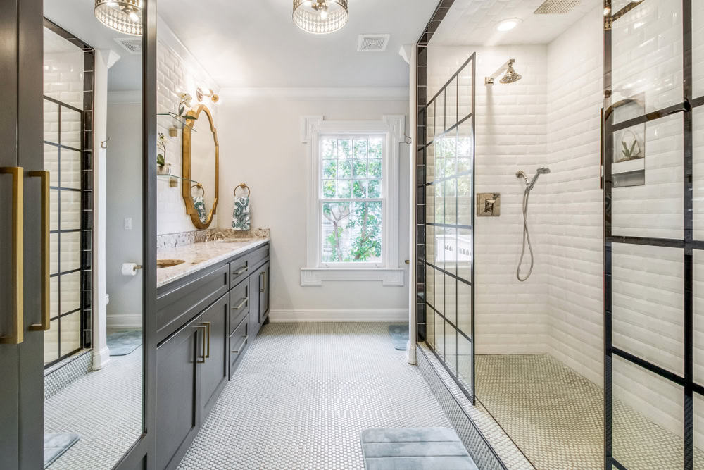 Modern bathroom featuring a long vanity with double sinks, round mirrors, elegant lighting, and a spacious walk-in shower enclosed with glass panels. The room is bright, with a window providing natural light and a patterned floor.