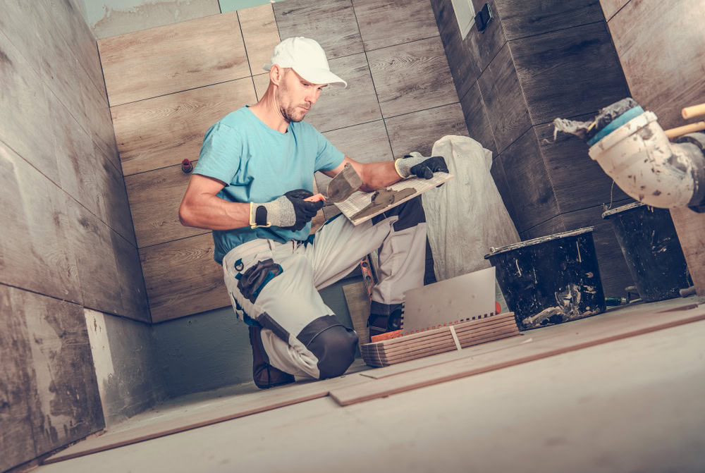 A construction worker kneels on the floor of a partially tiled room, cutting a tile with a tool. He wears a white cap, blue shirt, work gloves, and kneepads. Various tools and materials are scattered around him.