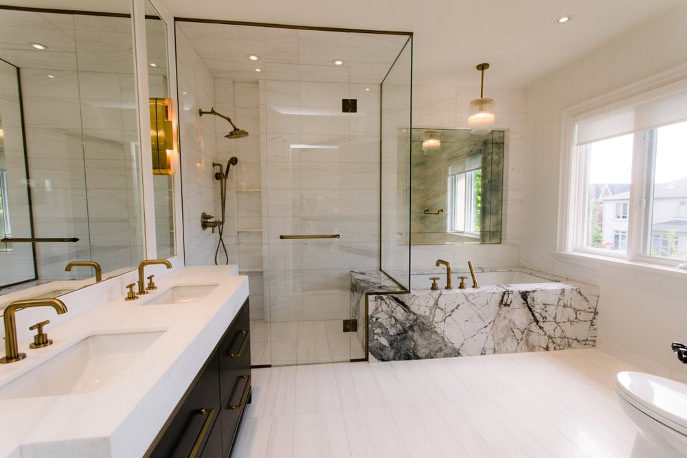 Modern bathroom with white marble and gold accents. Features include a double sink vanity, a spacious glass shower, a marble bathtub, large windows, and sleek lighting. The space is bright and elegant.