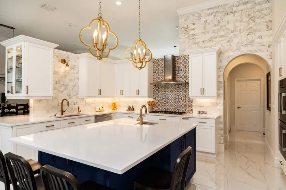 A modern kitchen featuring a large, white island with a dark blue base and gold fixtures. White cabinets and marble-patterned walls complement the patterned backsplash. Two elegant pendant lights hang above the island, and black chairs surround it.