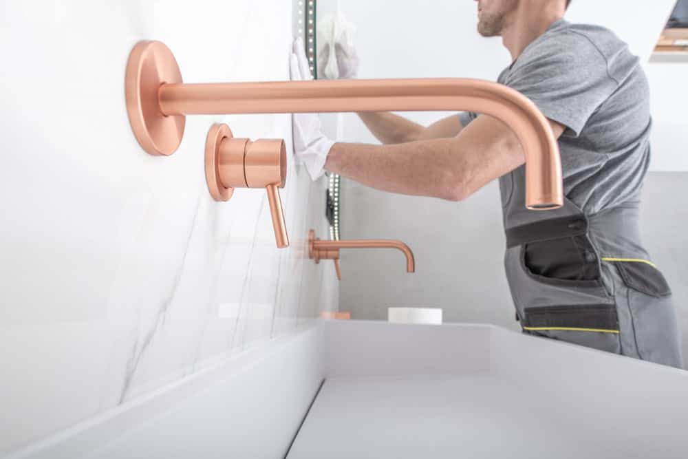 A person in a gray shirt and work apron is installing a mirror in a modern bathroom. The bathroom features a white sink and copper-colored wall-mounted faucet and handles.