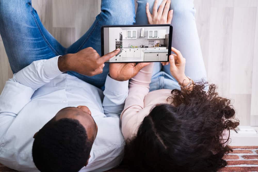 A couple lies on a wooden floor, looking at a tablet showing a kitchen design. The man points at the screen, showcasing white cabinets and a modern layout. They both wear casual clothing, with jeans visible.