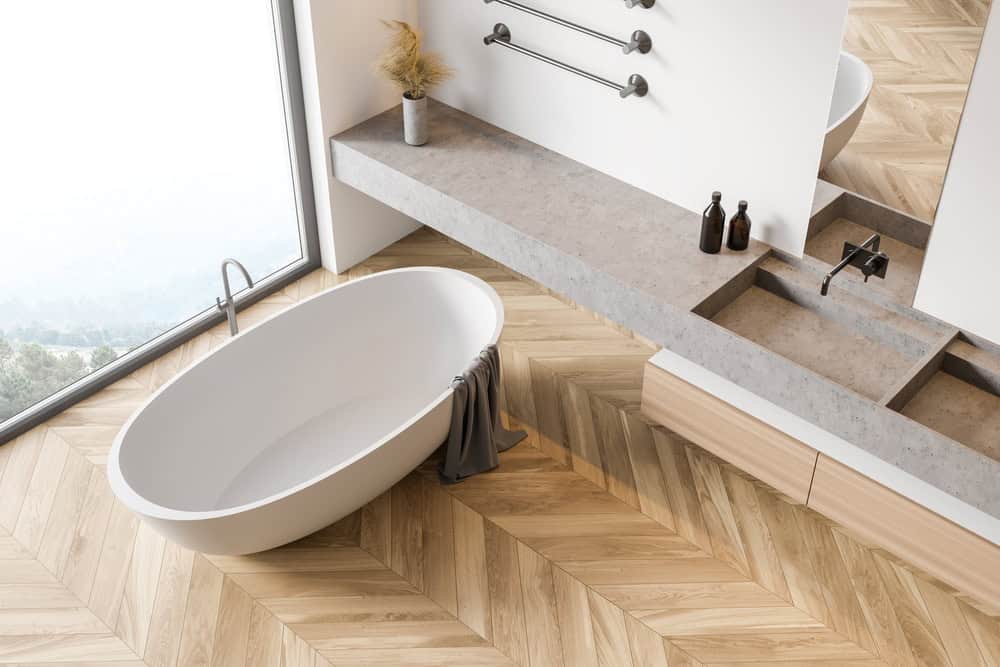 A modern bathroom with a freestanding oval bathtub on a wooden herringbone-pattern floor. A gray towel drapes over the tub's edge. Nearby, a stone countertop with dual sinks and black faucets, plus decorative bottles, enhances the minimalist decor.