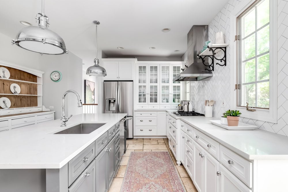 A bright, modern kitchen with white cabinets, stainless steel appliances, and gray countertops. Two pendant lights hang over a large island with a sink. A window on the right lets in natural light, highlighting a potted plant and decorative shelves.