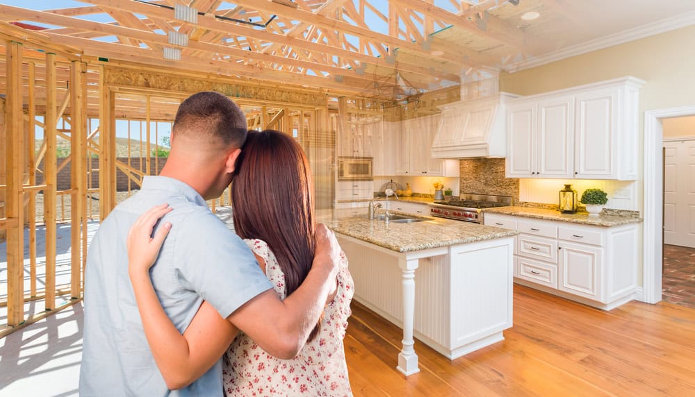 A couple with their backs to the camera embraces while looking at the inside of a house. The image blends a view of an unfinished wooden frame with a finished kitchen, featuring white cabinets and a granite countertop.