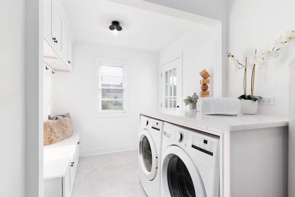 A bright, modern laundry room with white walls and cabinets showcases expert reconstruction services. A washing machine and dryer sit side by side under a countertop. A bench with a beige cushion complements the space, while natural light pours through the window onto a plant and neatly folded towels.