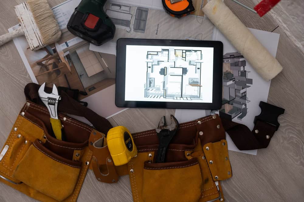 An overhead view of a tablet displaying a floor plan, surrounded by renovation tools including a wrench, pliers, measuring tape, paint roller, and a brush. A tan tool belt and design sketches are also visible on a wooden surface.