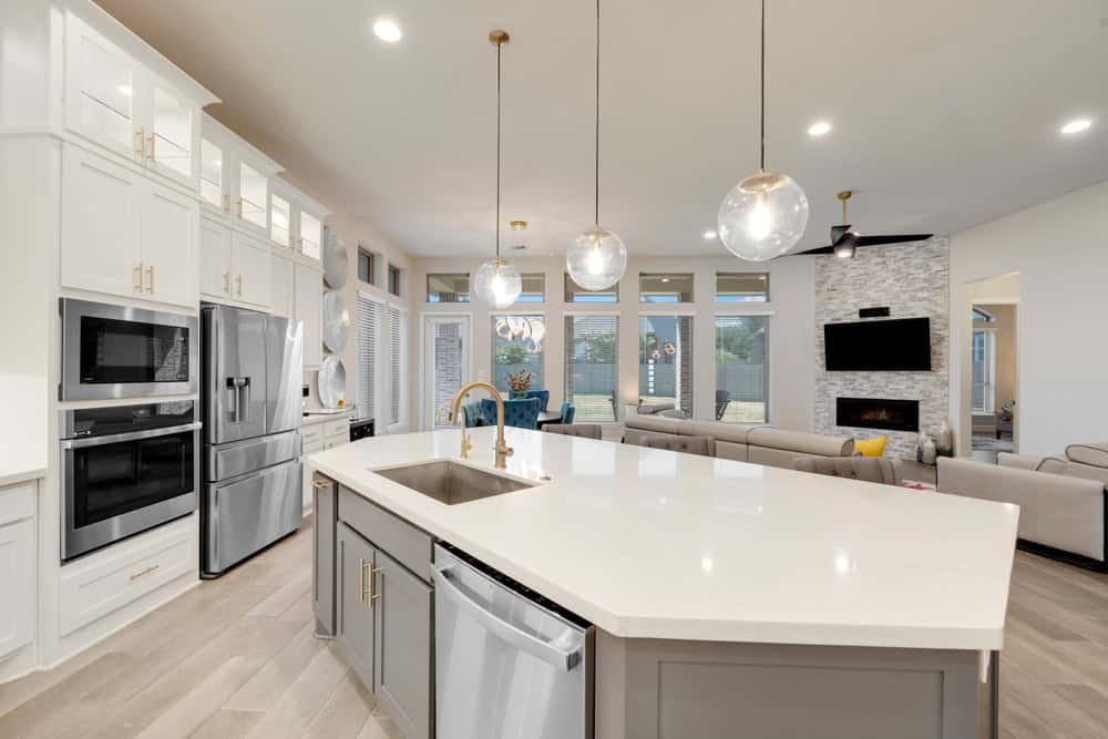 Modern kitchen and living area featuring a large island with a sink, elegant pendant lights, stainless steel appliances, and a gray and white color scheme. Recently enhanced with premium reconstruction services, the open-concept space leads to a cozy living room with a fireplace and large windows.