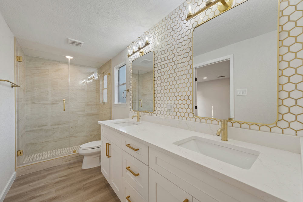 Modern bathroom featuring a double vanity with white countertops and gold accents. A large mirror with gold hexagonal tile backsplash is above the sinks, showcasing exquisite reconstruction services. A glass-enclosed shower is in the background, and the floor is light wood.